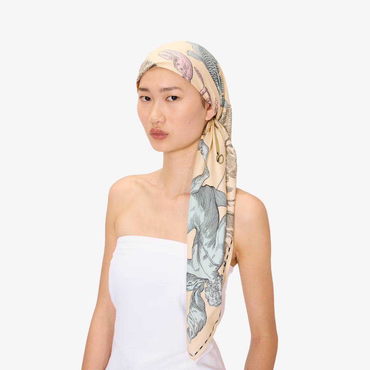 A person with long dark hair wears the INOUI EDITIONS Astrologie Scarf—a silk modal blend adorned with intricate zodiac signs—over a white strapless top, set against a plain white background.