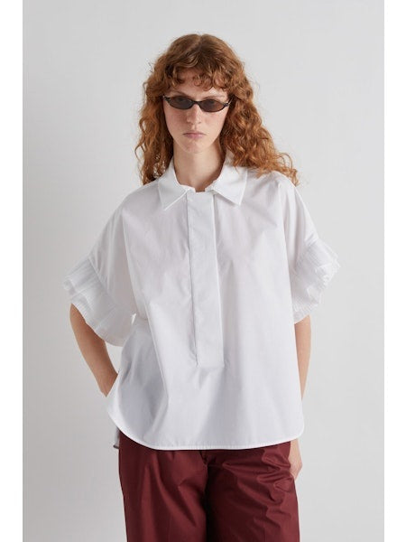 A person with long, wavy hair and sunglasses models the MEIMEIJ - WHITE COTTON SHIRT, featuring ruffled sleeves, paired with maroon pants, standing in profile against a plain white background.