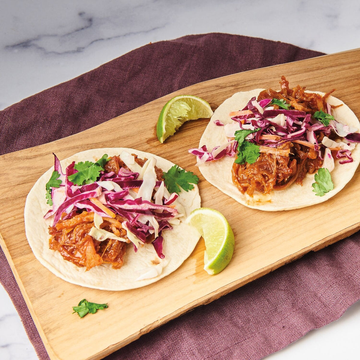 Two tortillas topped with shredded meat, red and green cabbage slaw, and cilantro sit on a wooden board with lime wedges on the side, placed on a purple cloth. A drizzle of DIVINA - CALABRIAN CHILI ORANGE SPREAD by DIVINA adds a sweet and spicy kick, perfectly complementing the vibrant flavors.