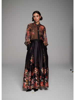 A woman poses in the MILVA MI - LONG FLORAL MAXI SKIRT by MILVA, paired with a sheer, long-sleeved floral blouse and pointed black heels against a simple studio backdrop.