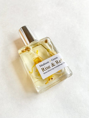 A small rectangular glass bottle labeled “MADISON AND GREEN - RISE & REFRESH MORNING AROMATHERAPY BODY OIL MINI” from MADISON GREEN, featuring a silver cap and visible botanical pieces suspended in the lightweight, plant-based oil.
