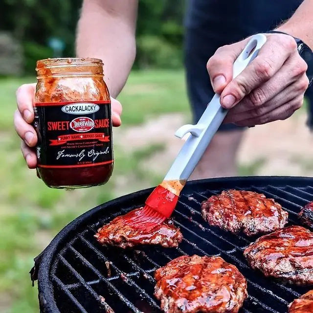 A person brushes savory barbecue sauce onto the burgers sizzling on the grill, holding a jar of CACKALACKY - CHEERWINE SWEET SAUCE in their other hand. This scene of Southern cuisine unfolds against the backdrop of a grassy outdoor setting.