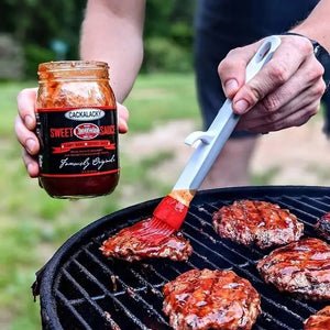 A person brushes savory barbecue sauce onto the burgers sizzling on the grill, holding a jar of CACKALACKY - CHEERWINE SWEET SAUCE in their other hand. This scene of Southern cuisine unfolds against the backdrop of a grassy outdoor setting.