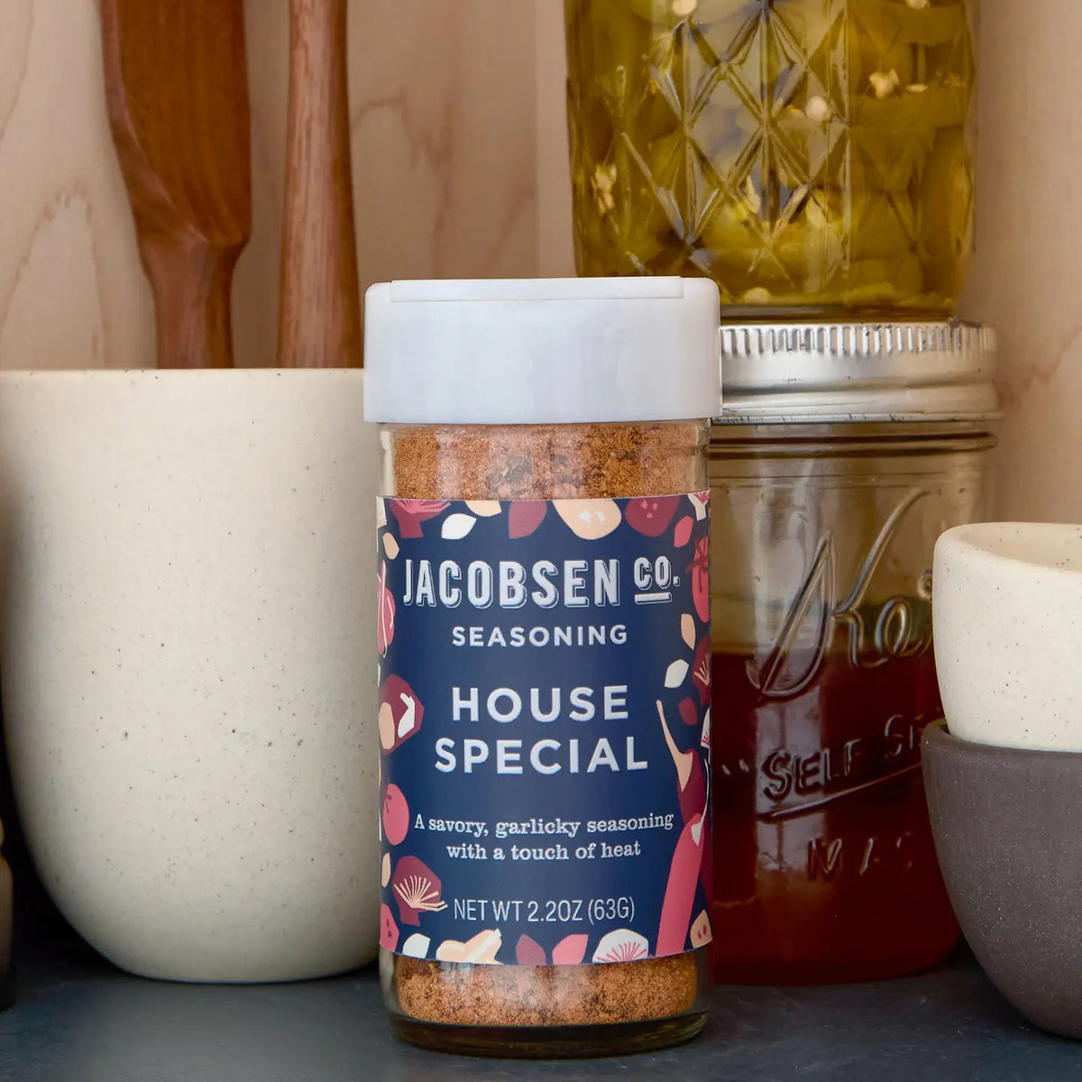 A jar of JACOBSEN SALT CO. HOUSE SPECIAL SEASONING sits on a kitchen counter, surrounded by a ceramic bowl, a glass jar of honey, a bottle of oil, and wooden utensils.