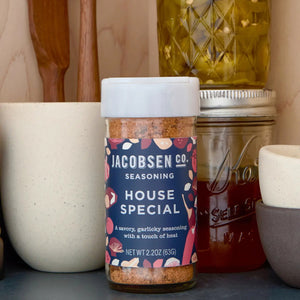 A jar of JACOBSEN SALT CO. HOUSE SPECIAL SEASONING sits on a kitchen counter, surrounded by a ceramic bowl, a glass jar of honey, a bottle of oil, and wooden utensils.