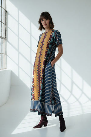 A woman wears the BRITON COURT - DIANA MAXI DRESS by BRITON COURT, a loose-fitting viscose maxi dress featuring vertical stripes in yellow, blue, and brown, short sleeves, practical side pockets, and an ankle-length hem. She is standing against a plain white background and pairs the dress with dark ankle boots.