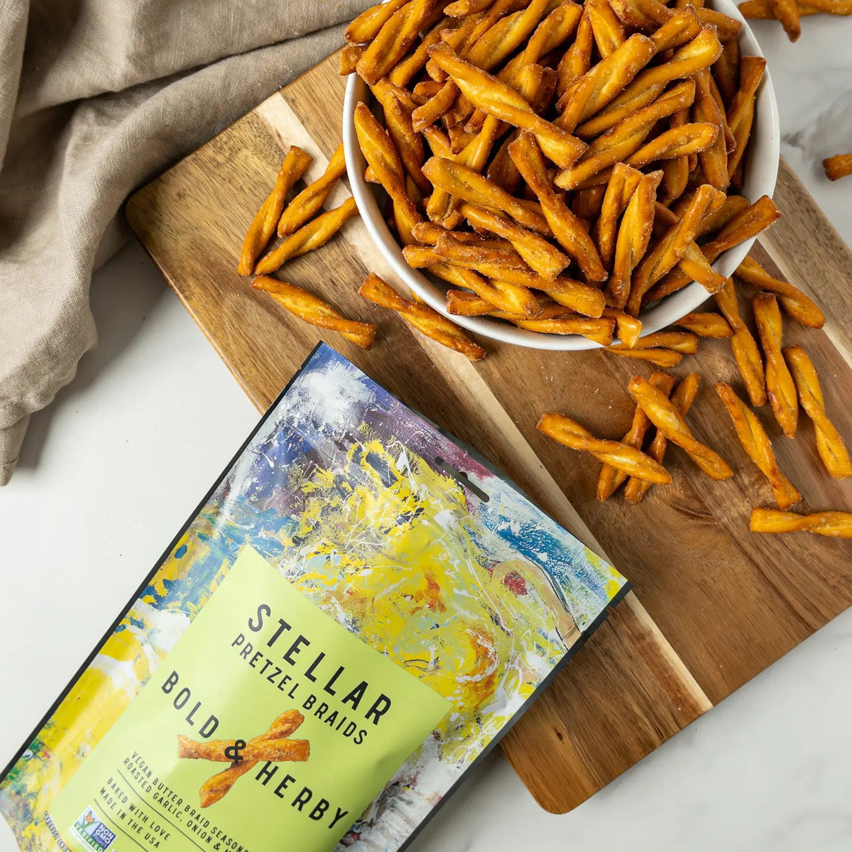 A vibrant bag of STELLAR SNACKS - BOLD AND HERBY PRETZEL BRAIDS, vegan pretzel twists flavored with roasted garlic and onion, highlighted by a green label and abstract art design—an irresistibly tasty non-GMO snack from STELLAR SNACKS that’s perfect for any occasion.
