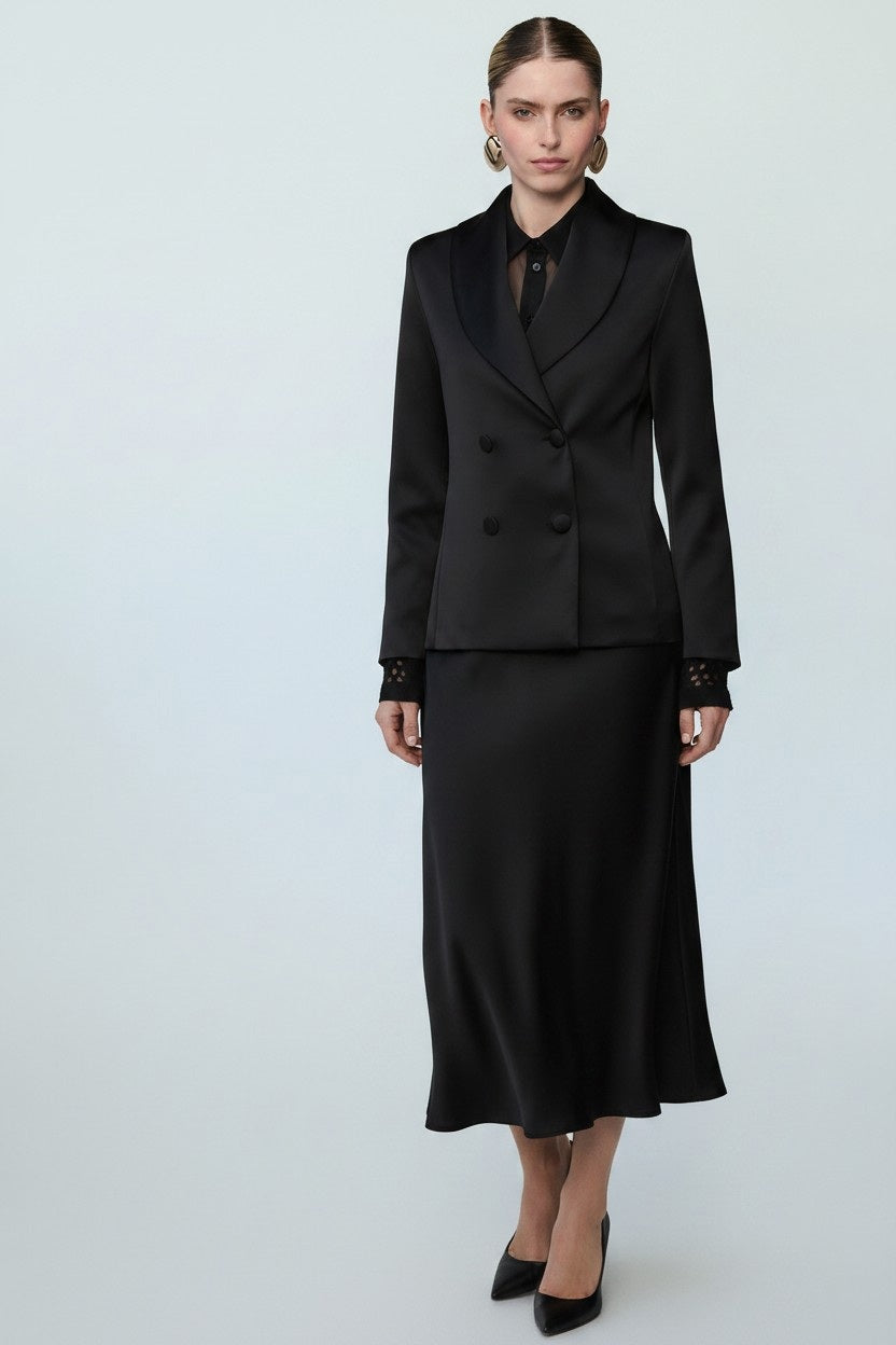 A woman stands against a plain light background wearing the MILVA MI - BLACK SATIN BLAZER by MILVA, paired with a matching long skirt, black blouse, black heels, and gold hoop earrings.