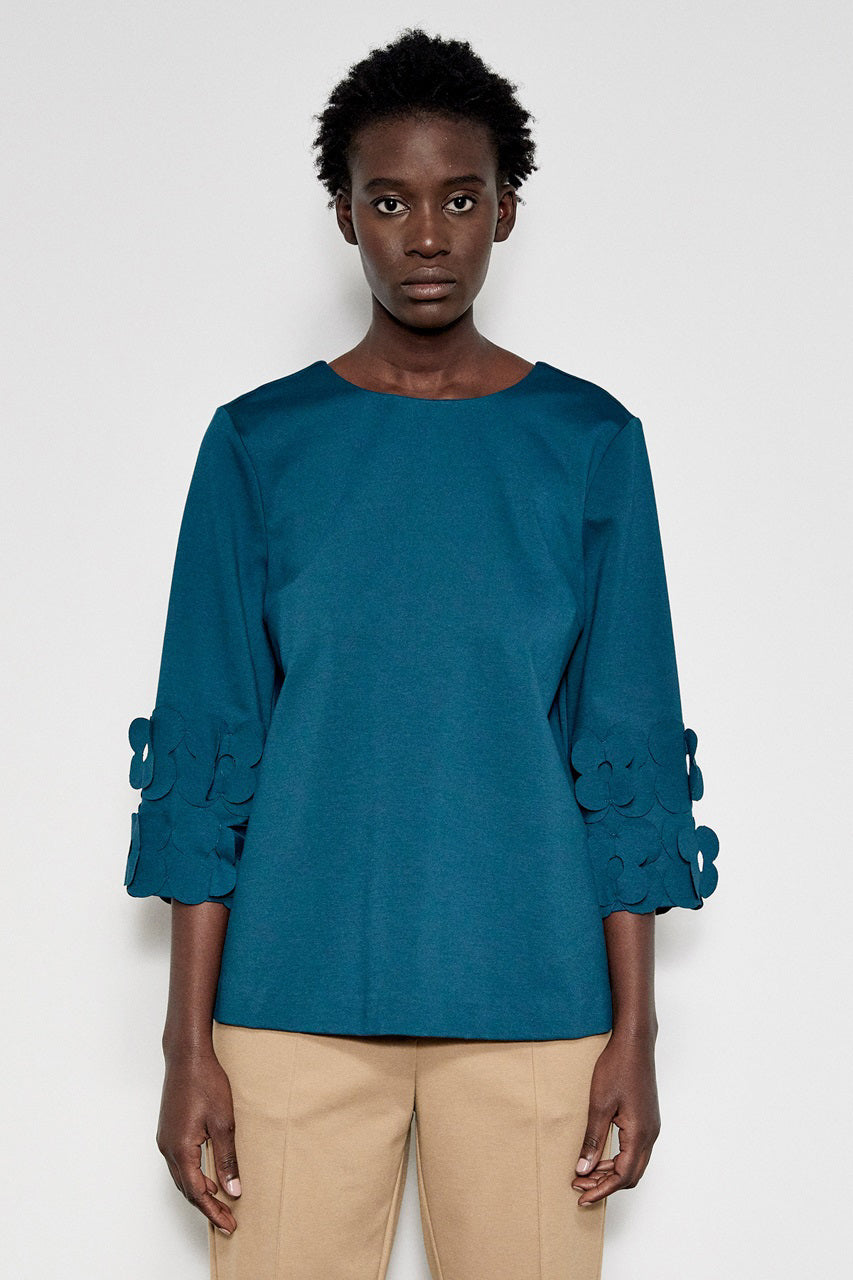 A person with short curly hair stands in profile against a white background. They are wearing the MEIMEIJ - 3/4 Sleeve Back to Front Top with Lasercut Flower Detail, a loose-fitting teal top featuring flower embellishments on the sleeves, paired with light beige pants.