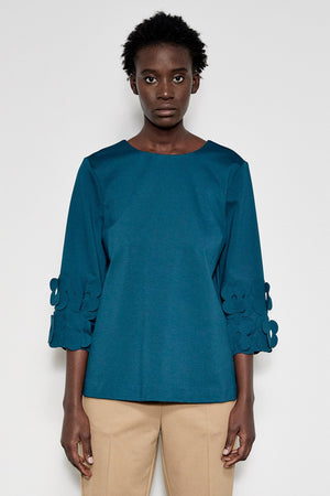 A person with short curly hair stands in profile against a white background. They are wearing the MEIMEIJ - 3/4 Sleeve Back to Front Top with Lasercut Flower Detail, a loose-fitting teal top featuring flower embellishments on the sleeves, paired with light beige pants.