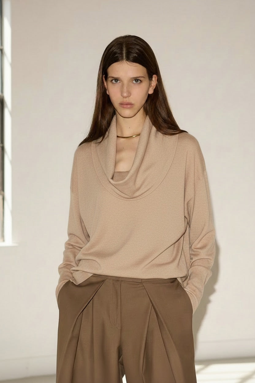 A woman with long brown hair wears the NUDE Draped Cowl Neck Sweater in virgin wool off white, made in Italy, tucked into loose brown trousers as she stands in front of a white wall with a window on the left.