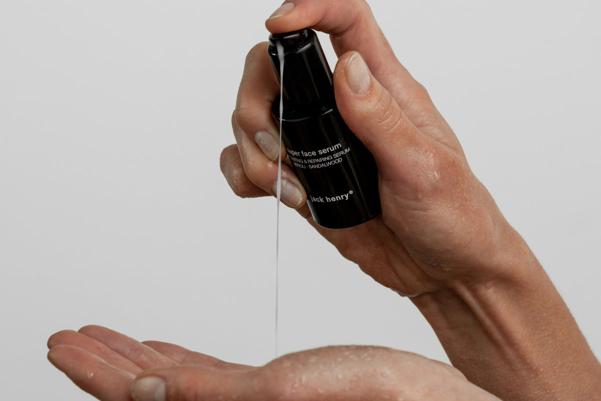 A person dispenses JACK HENRY - SUPER FACE SERUM, an anti-aging formula, from a black pump bottle into their hand against a plain white background.