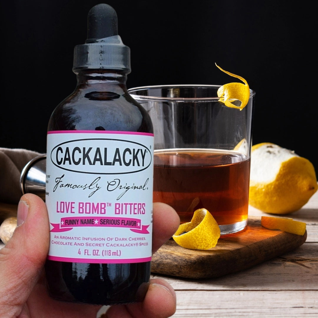A 4 oz. dark glass dropper bottle of CACKALACKY - LOVE BOMB BITTERS from CACKALACKY, featuring pink and white text on the label, sits against a plain white background—an ideal addition to your aromatic cocktail bitters collection.