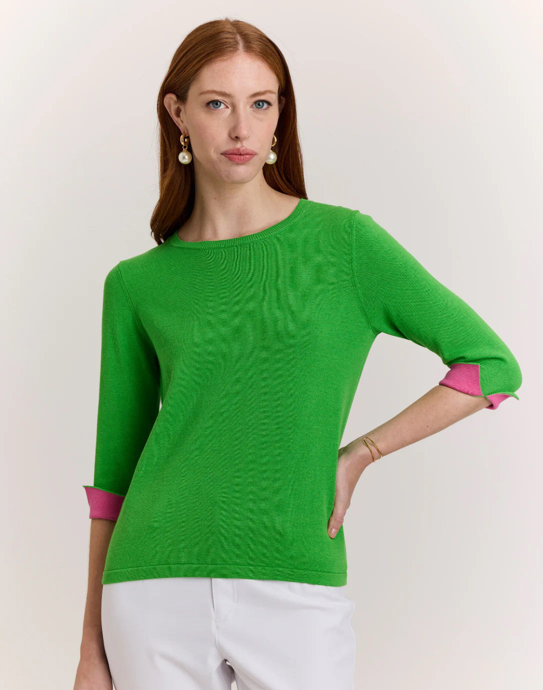 A woman with long red hair models the HINSON WU Tyler 3/4 Sleeve Knit Sweater in bright green with pink cuffs, paired with white pants, standing against a plain light background.