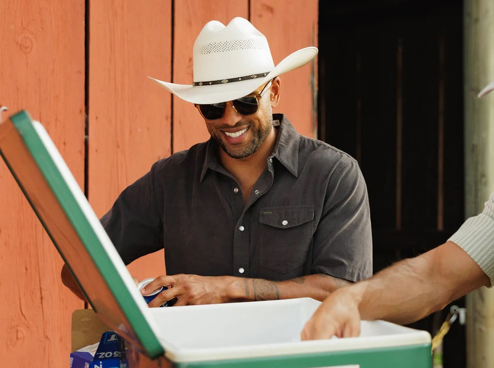 A smiling man in a brown hat and the CRIQUET Short Sleeve Corduroy Pearl Snap Shirt in Iron Horse holds a canned drink in his right hand, standing against a white background.