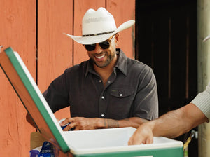 A smiling man in a brown hat and the CRIQUET Short Sleeve Corduroy Pearl Snap Shirt in Iron Horse holds a canned drink in his right hand, standing against a white background.