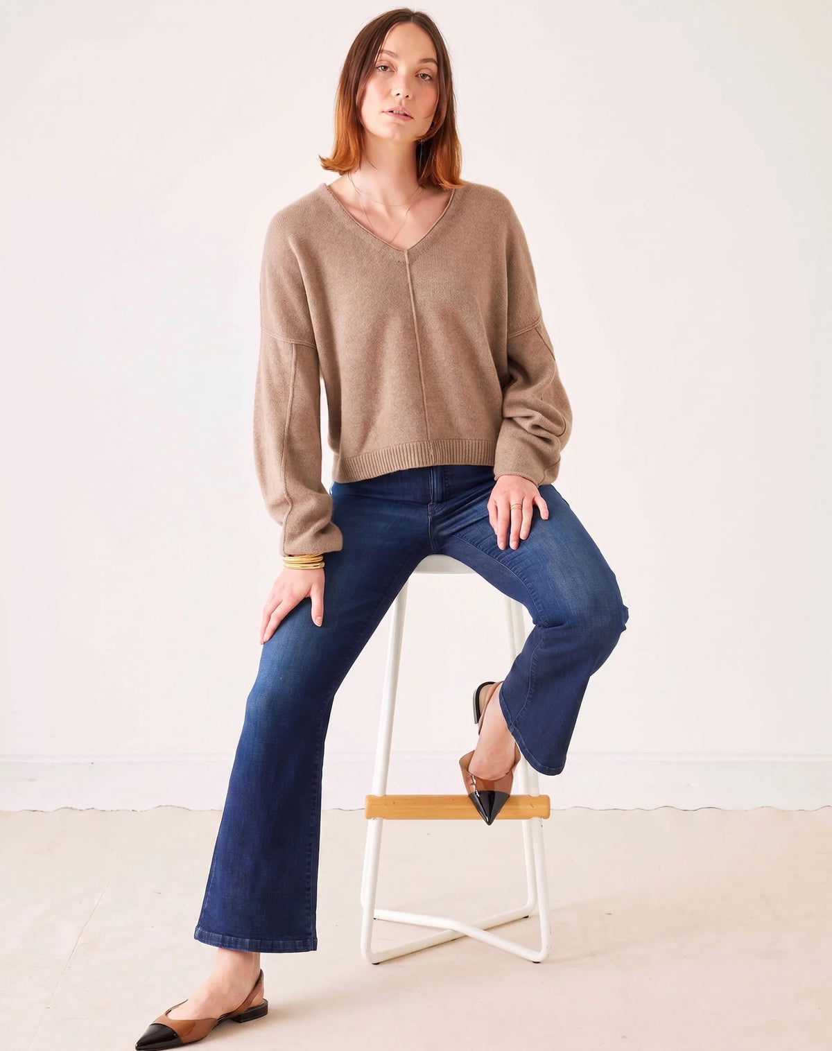 A person stands with hands in pockets against a plain light background, wearing MERSEA’s Murphy Mini Boot Jeans in Patriot—a pair of mid-rise, blue flare jeans crafted from four-way stretch denim—paired with a tan long-sleeve top, black pointed-toe flats, and gold bangles.