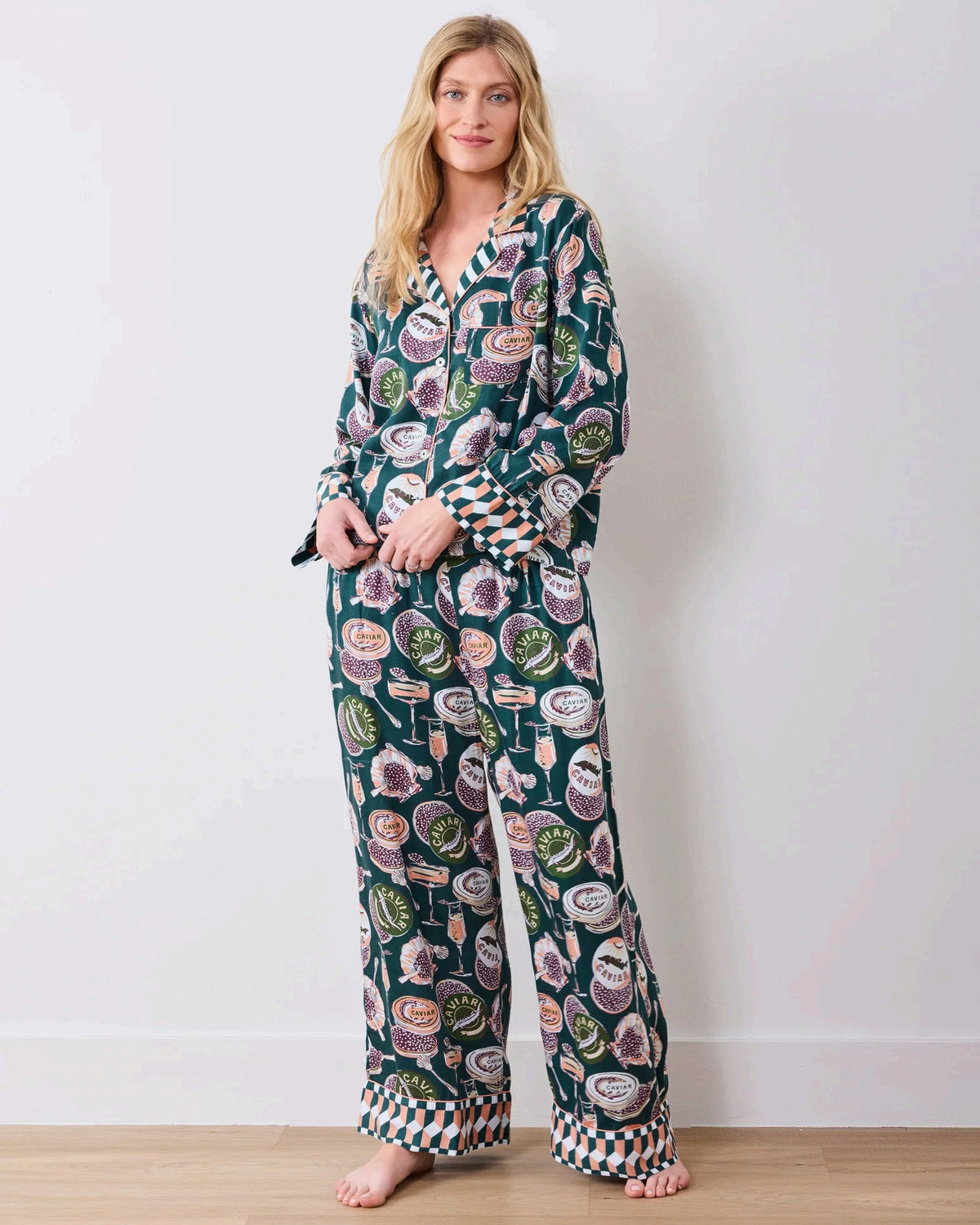 A woman stands barefoot on a wooden floor against a white wall, wearing the PRINTFRESH - Champagne & Caviar Eco Satin Soho Set in Peacock Feather by PRINTFRESH, a matching dark loungewear set adorned with colorful floral and fruit motifs and finished with checkered cuffs.