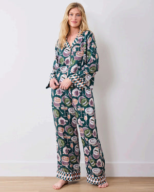 A woman stands barefoot on a wooden floor against a white wall, wearing the PRINTFRESH - Champagne & Caviar Eco Satin Soho Set in Peacock Feather by PRINTFRESH, a matching dark loungewear set adorned with colorful floral and fruit motifs and finished with checkered cuffs.