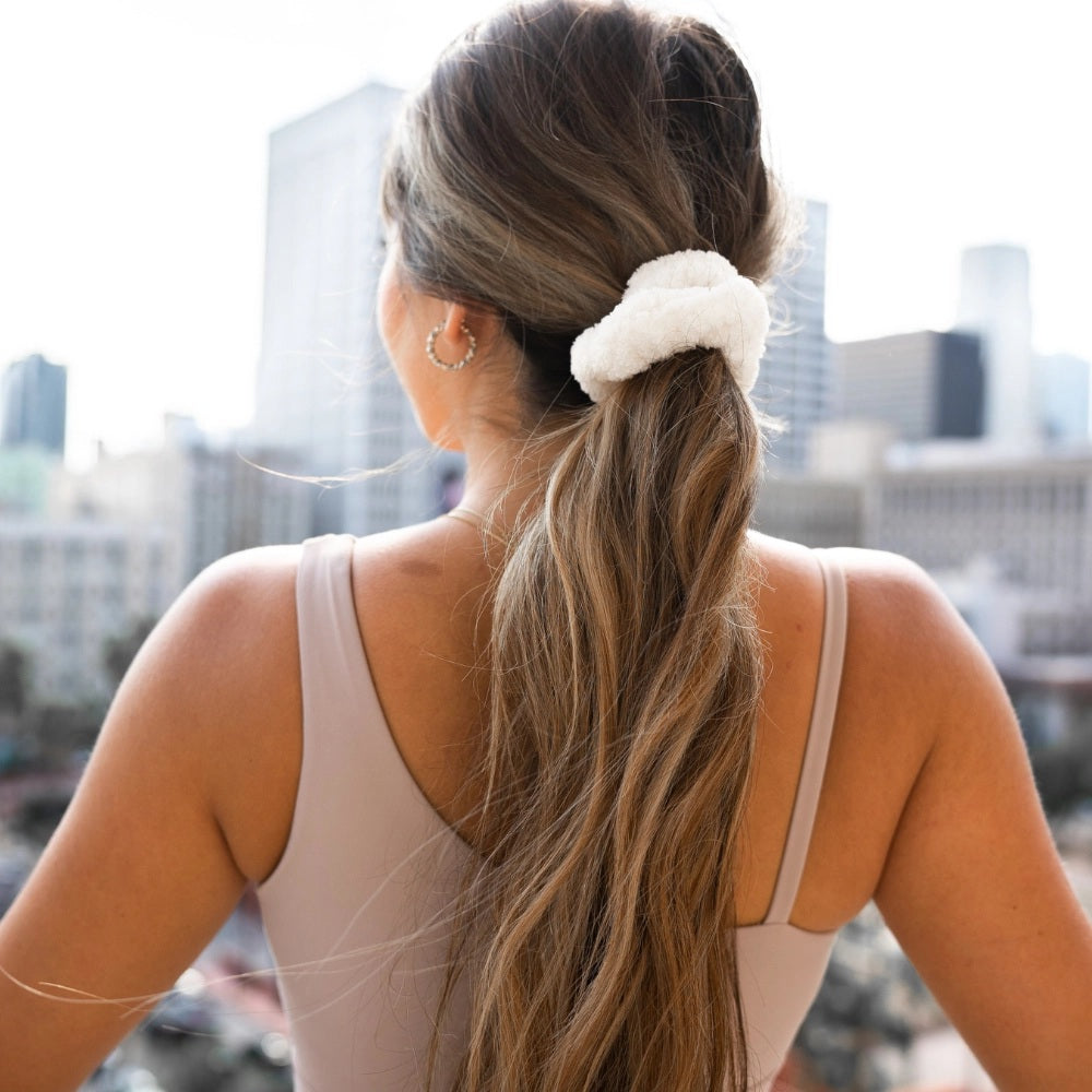 A woman with wavy, light brown hair in a ponytail secured with a VOLO BEAUTY - SCRUNCHIE DUO PACK is shown from the back, wearing a black shirt and gold jewelry, and raising her left hand.