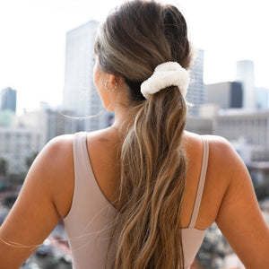 A woman with wavy, light brown hair in a ponytail secured with a VOLO BEAUTY - SCRUNCHIE DUO PACK is shown from the back, wearing a black shirt and gold jewelry, and raising her left hand.