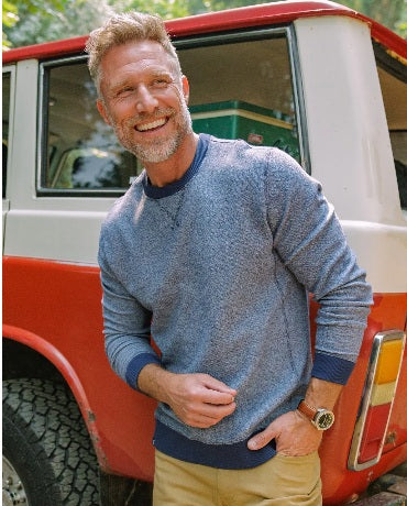A man with short brown hair wearing the CRIQUET - French Terry Crewneck Sweatshirt in Dusty Blue and white pants stands with one hand in his pocket, looking to his left against a white background.