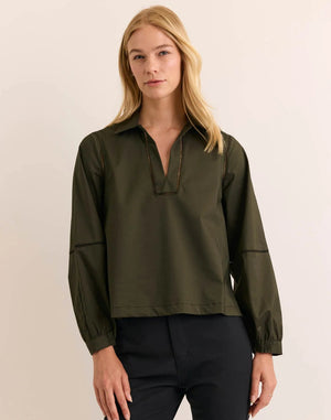 A woman with long blonde hair stands against a plain light background, wearing the HINSON WU LAURA Long Sleeve Lace Trim Top in Oregano—a refined, machine-washable blouse featuring a collared neckline—paired with black pants.