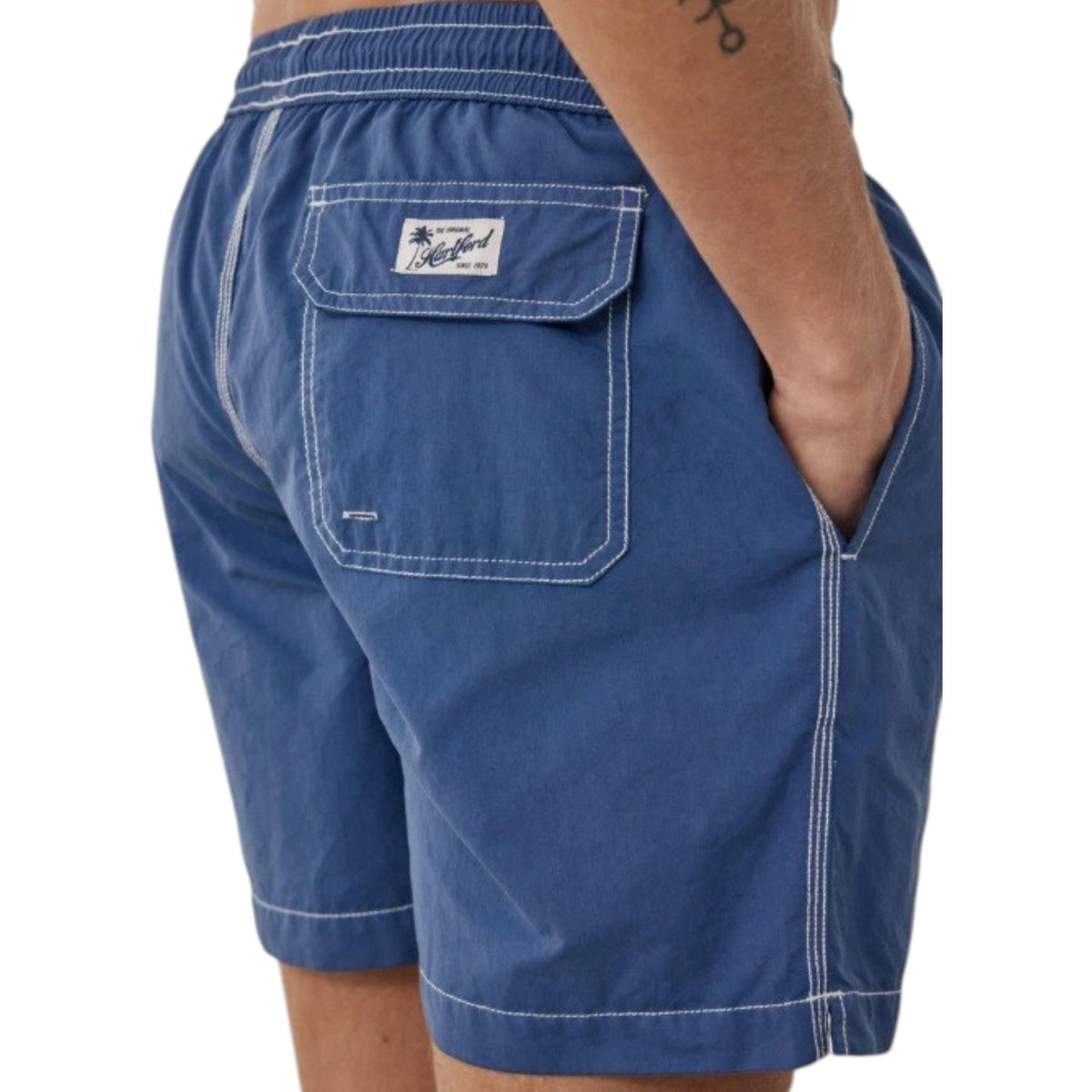 A shirtless person models the HARTFORD - NYLON SWIM TRUNKS WITH TRAVEL POUCH IN DENIM by HARTFORD against a plain light background, with tattoos visible on their arms and legs and one hand casually in a pocket.