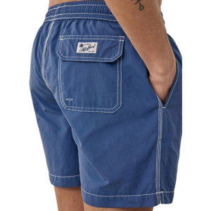 A shirtless person models the HARTFORD - NYLON SWIM TRUNKS WITH TRAVEL POUCH IN DENIM by HARTFORD against a plain light background, with tattoos visible on their arms and legs and one hand casually in a pocket.
