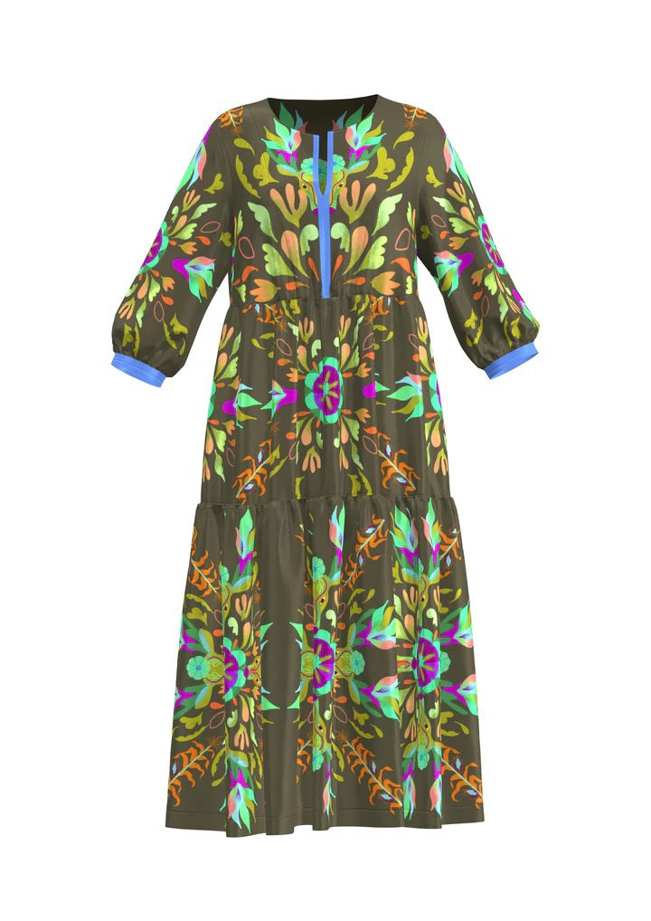 A woman wears the FRANCO FERRARI - SILK TIERED MIDI DRESS, a long sleeve olive green silk piece with colorful floral and abstract prints. She smiles softly, has wavy hair, beige sandals, and stands against a plain light gray background.