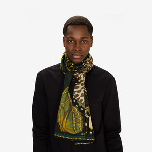 A woman in a light gray sweater and black pants stands against a plain white background, wearing the INOUI EDITIONS - PAPILLON (BUTTERFLIES) wool scarf in emerald from INOUI, featuring black and yellow butterfly designs.
