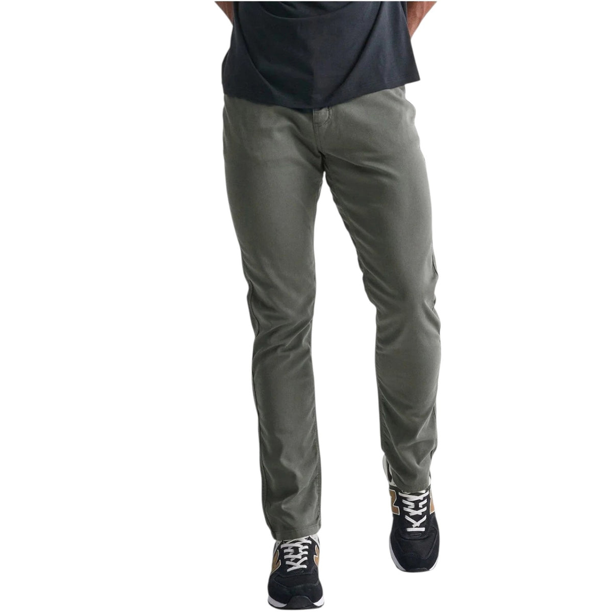 A person stands against a plain white background wearing DUER No Sweat Relaxed Taper Pants in Gull (olive green), a black T-shirt, and black New Balance sneakers with white soles and gold accents.