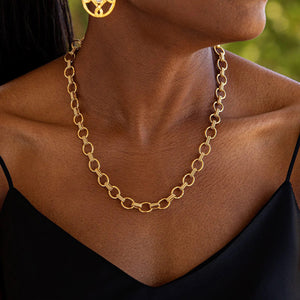 A person adorned with a CAPUCINE DE WULF - CLEOPATRA SMALL LINK NECKLACE featuring delicate interlocking round links and a pair of gold hoop earrings. The individual is dressed in a black sleeveless top, standing against the backdrop of a natural, blurred green setting.
