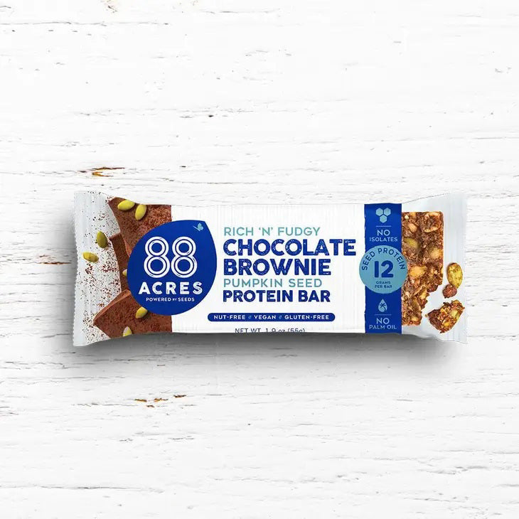A 88 Acres Dark Chocolate Brownie High Protein Bar wrapper rests on a white wooden surface. The predominantly white and blue wrapper features the brand name &quot;88 Acres&quot; alongside the product name &quot;Rich &#39;n&#39; Fudgy Dark Chocolate Brownie Pumpkin Seed High Protein Bar.&quot; Text highlights that it is nut-free, vegan, and gluten-free.