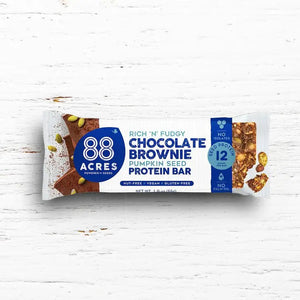 An 88 ACRES - DARK CHOCOLATE BROWNIE HIGH PROTEIN BAR sits on a white wooden surface. The nut-free, vegan, and gluten-free packaging highlights it contains 12g of protein.