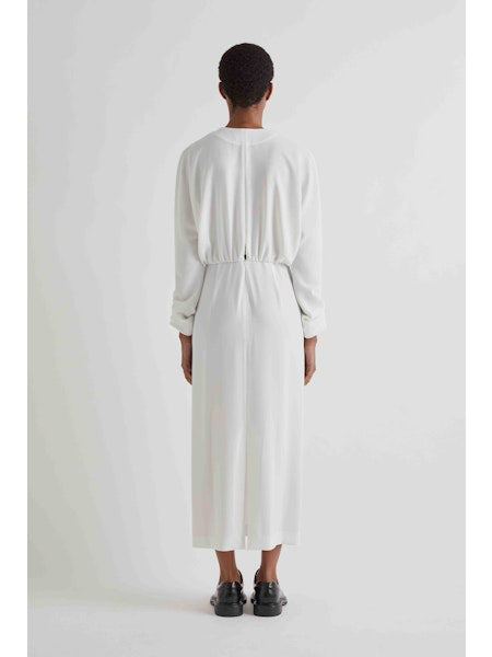 A person in profile wears the MEIMEIJ Gathered Waist Long Sleeve Dress in white, paired with black shoes and sunglasses. They have short hair and stand against a plain light background.