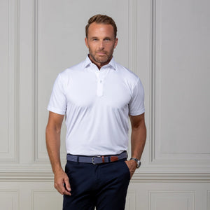 A man in sunglasses and a white COLLARS AND CO - FEATHERWEIGHT FLEX POLO stands outdoors, smiling and holding his collar, with greenery and a stone wall in the background.