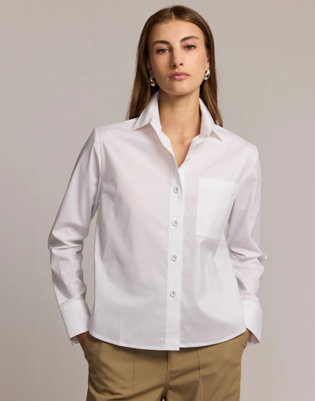 A woman models the HINSON WU - VENUS LONG SLEEVE UNTUCKABLE SHIRT in white stretch polished cotton with a chest pocket, paired with beige pants, standing against a neutral background and holding the shirt collar.