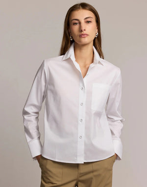 A woman models the HINSON WU - VENUS LONG SLEEVE UNTUCKABLE SHIRT in white stretch polished cotton with a chest pocket, paired with beige pants, standing against a neutral background and holding the shirt collar.
