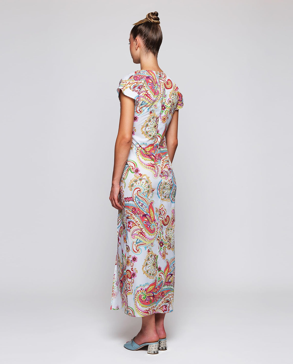 A woman stands against a white background wearing the MIRTO 1956 - PAISLEY PRINTED MIDI DRESS with short sleeves and light blue open-toe heels, her hair in a high bun.