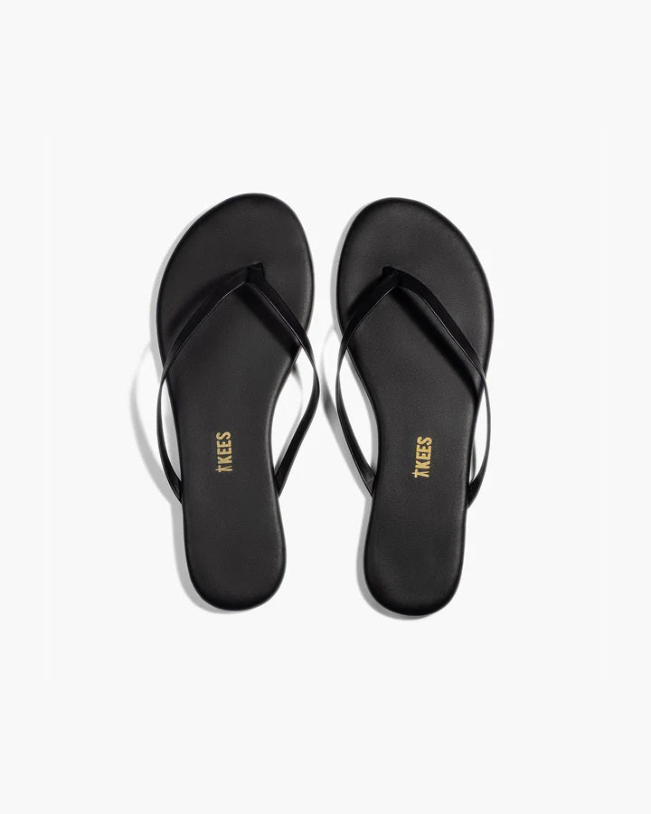 A pair of TKEES - LILY SLINER SLIPPER SABLE by TKEES INC, featuring black flip-flop sandals with thin straps and “TKEES” in gold on the cushioned insole, shown on a white background.
