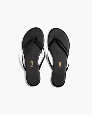 A pair of TKEES - LILY SLINER SLIPPER SABLE by TKEES INC, featuring black flip-flop sandals with thin straps and “TKEES” in gold on the cushioned insole, shown on a white background.