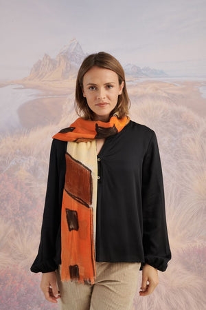 A woman in a black long-sleeve top and beige pants stands before a scenic backdrop, her look elevated by the STORIATIPIC ISCHIA Modal and Wool Scarf (100x200 cm) in vibrant orange and brown, draped elegantly around her neck.