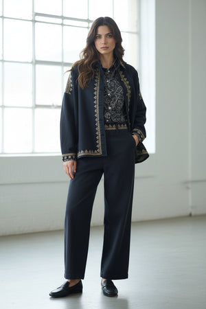 A woman with long brown hair stands indoors in front of large windows, embodying effortless style in the STORIATIPIC ROXY JACKET IN NOIR by STORIATIPIC, featuring gold embroidery on 100% cotton. She pairs the jacket with a patterned shirt, dark pants, and black loafers, mindful to follow proper cotton care such as gentle machine wash.