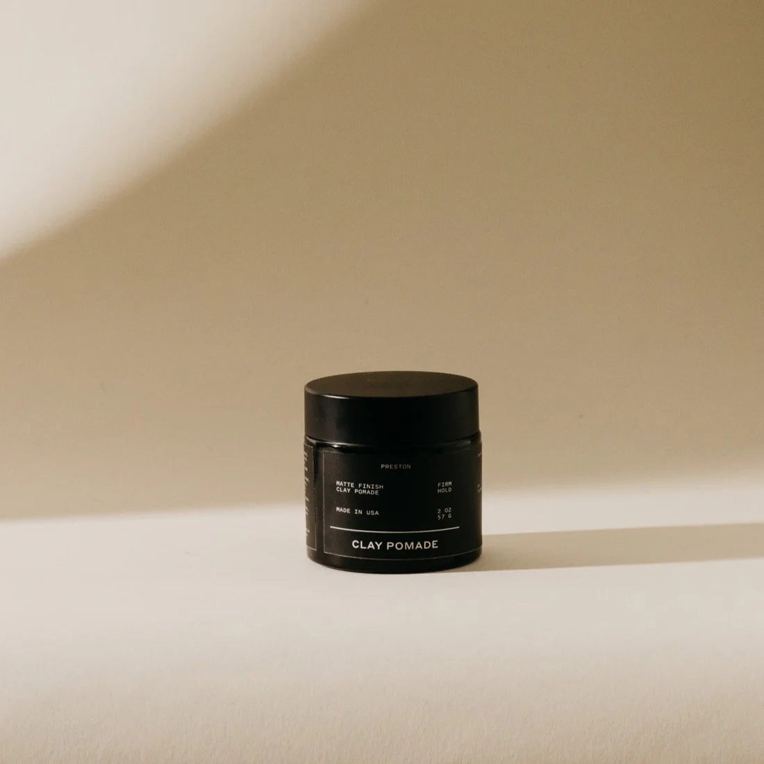 A small, dark-colored jar of PRESTON - CLAY POMADE UNSCENTED sits on a neutral-colored surface, casting a shadow. Text on the jar reads "PRESTON," "CLAY POMADE," "MATTE FINISH," "HIGH HOLD," "MADE IN USA," and other smaller details. Ideal for medium to thicker hair types, the background is softly lit, creating a minimalist look.