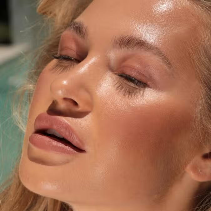 A close-up of a person with curly dark hair, a serious expression, and warm makeup featuring the universally flattering, weightless JASON WU - HOT FLUFF LIP/EYE/CHEEK by JASON WU BEAUTY on their face. They are looking directly at the camera with a neutral background.