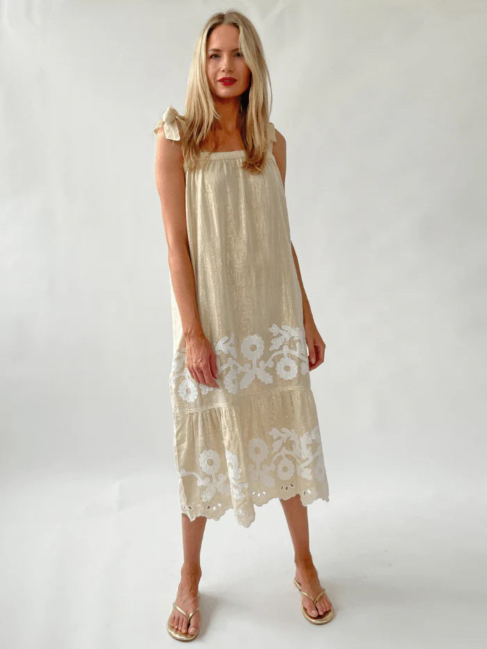 A woman with long blonde hair wears the ROSE &amp; ROSE - ALBA SUNDRESS, a sleeveless beige maxi adorned with daisy appliqués and white floral embroidery at the hem. Crafted from a cotton gold lurex blend, she stands barefoot on a neutral background, gazing directly at the camera with a relaxed expression.