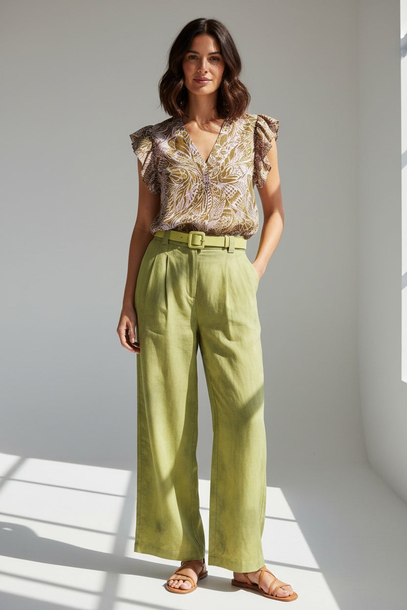 A woman with wavy blonde hair stands outdoors in a patterned SILK PRINTED BLOUSE by MOMONI and high-waisted green pants with a matching belt, gazing at the camera with a relaxed expression amid lush greenery.