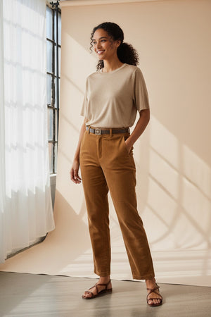 A woman with long brown hair wears the MOMONI - IORA LUREX JERSEY T SHIRT in gold, paired with beige and blue wide-leg floral pants and brown sneakers, standing against a neutral studio background.