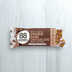 An 88 ACRES - DOUBLE DARK CHOCOLATE SEED + OAT BAR rests on a light wooden surface. The packaging is brown and white, showcasing images of dark chocolate chunks and oats. The label proudly highlights that it is gluten-free, vegan, and nut-free.